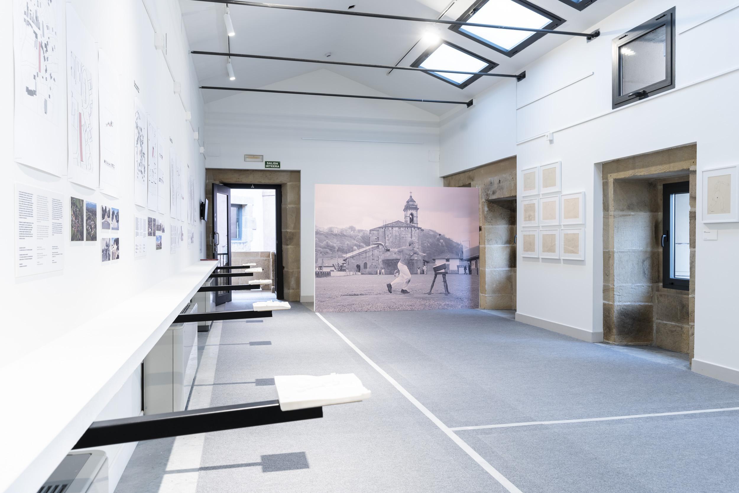 En esta imagen se puede ver la exposición de Pilotalekuak. En una sala grande con las paredes con expositores y al fondo una imagen antigua de un hombre con una cesta en la mano.